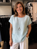 A woman with shoulder-length brown hair, wearing the ETERNELLE Top Sky Blue 6-SS26237-9 and white pants, smiles in a clothing store with racks of clothes visible in the background.