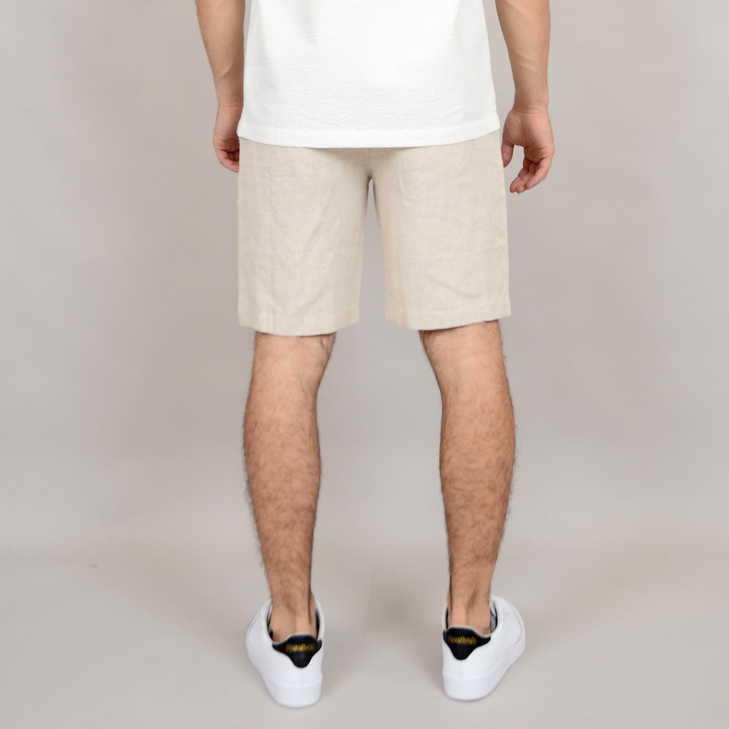 A person stands with their back to the camera against a plain light background, wearing a white shirt, white sneakers with black detail, and HEDGE Linen Blend Shorts Natural 41MW507S.