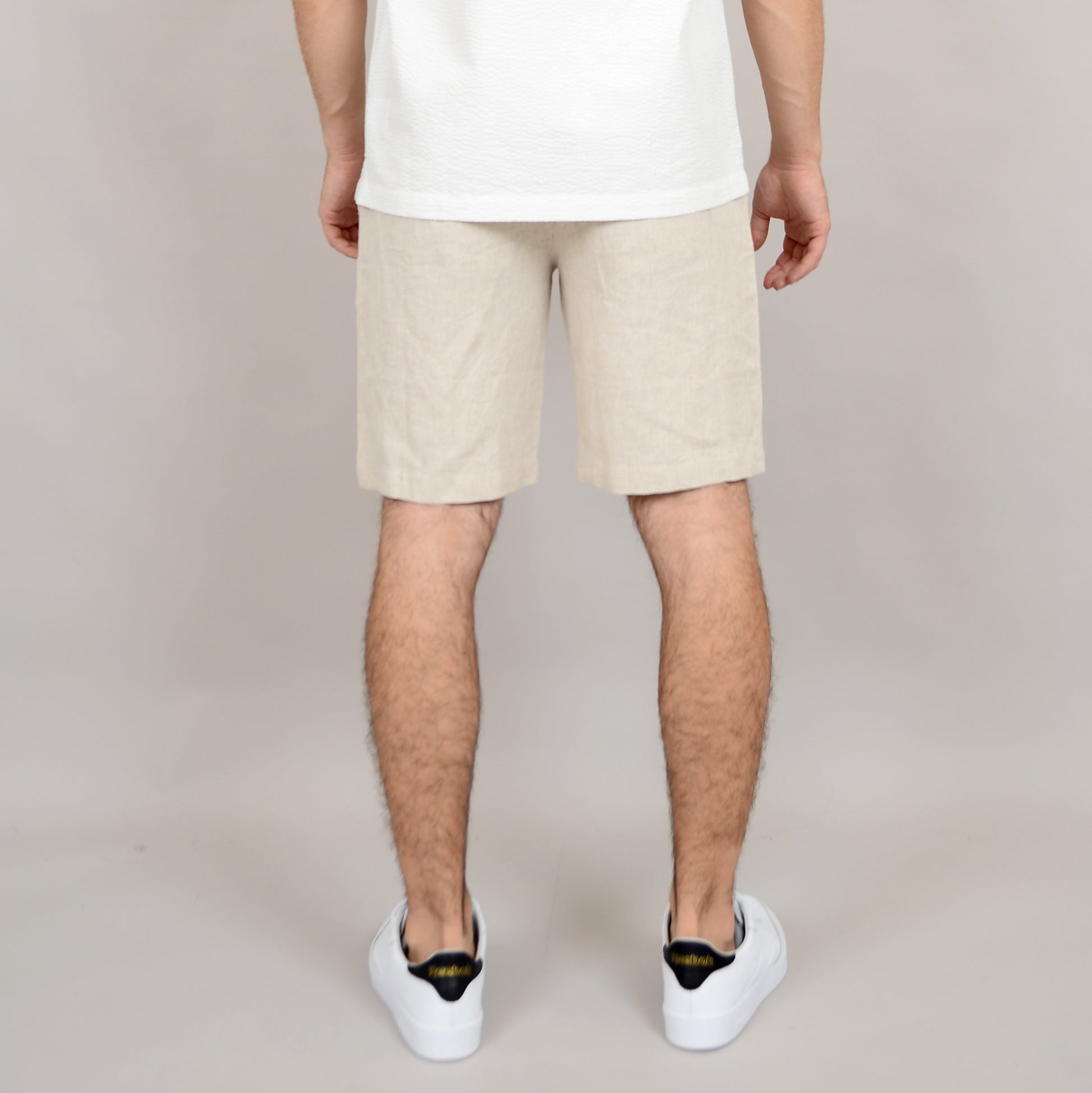 A person stands with their back to the camera against a plain light background, wearing a white shirt, white sneakers with black detail, and HEDGE Linen Blend Shorts Natural 41MW507S.