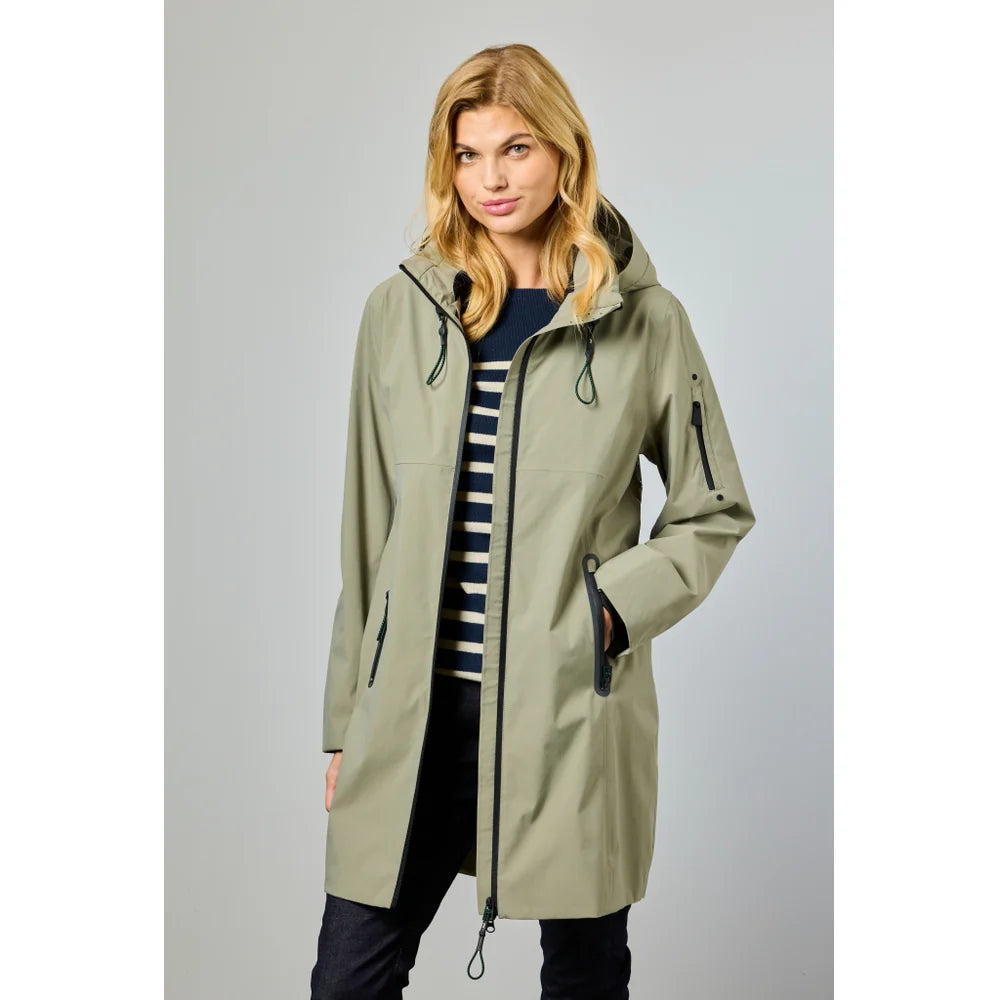 A woman with blonde hair wears the JUNGE Nynne Jacket in light olive green with reflective prints, layered over a navy and white striped sweater and dark pants, standing against a plain light gray background.