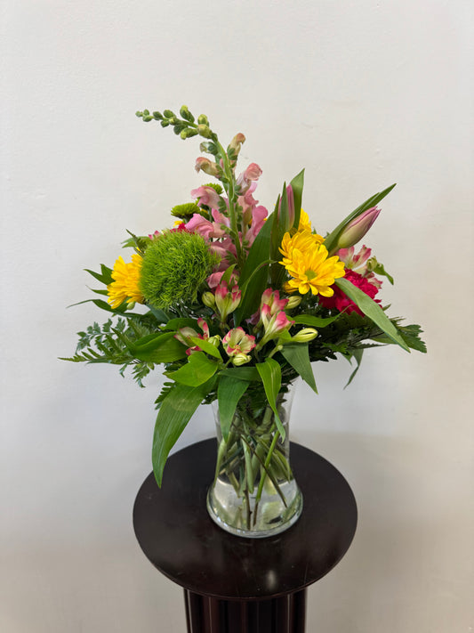 The Bright Moments Vase by Posies Flowers & Fashion displays a vibrant mix of blooms and greenery in a clear glass vase; flower varieties may vary based on availability.