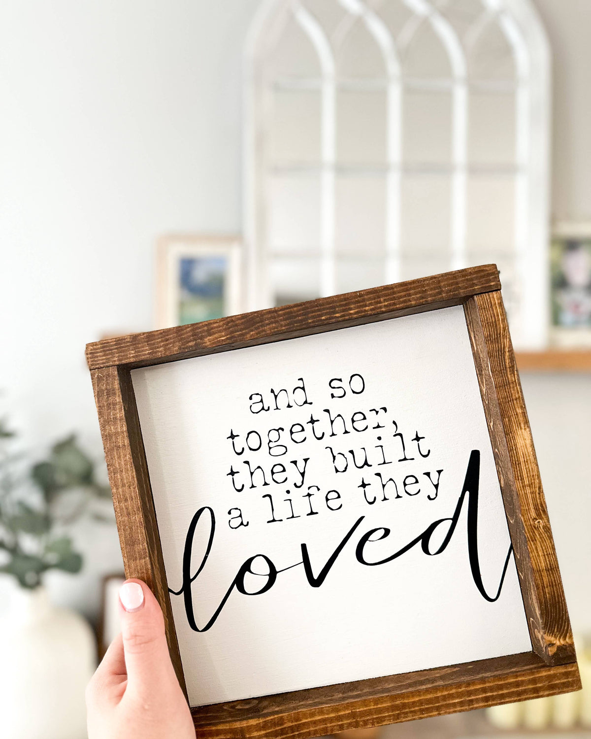 A hand holds a framed wood sign from Inspired Findings, featuring the phrase “and so together, they built a life they loved” in decorative script; the blurred home decor background highlights the warmth of handmade pieces like this “Built a life they loved (sm)” sign.