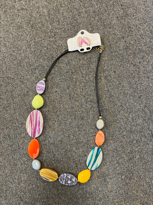 The Rainbow Road Necklace and Earrings set by Posies Flowers & Fashion Inc. lies on a grey surface, showcasing its bright and colorful oval-shaped beads with vibrant patterns in purple, pink, yellow, orange, and green. The necklace features an adjustable black cord and has a white tag attached to it. This stunning accessory pairs perfectly with the matching earrings for a complete look.