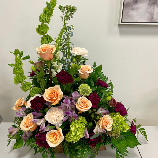 Introducing the Gentle Memories arrangement by Posies Flowers & Fashion—a lush funeral tribute featuring peach roses, green hydrangeas, purple alstroemeria, burgundy carnations, and white chrysanthemums. Accented with greenery, it creates a comforting atmosphere against a plain white background.