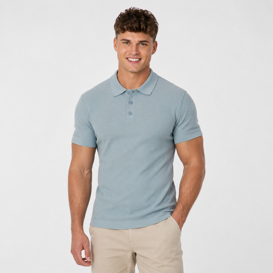 A young man with short brown hair smiles with one hand in his pocket, wearing beige pants and a RAGWEAR Polo Tarsh Short 2612-15050-2046 in Dusty Blue, standing against a plain white background.