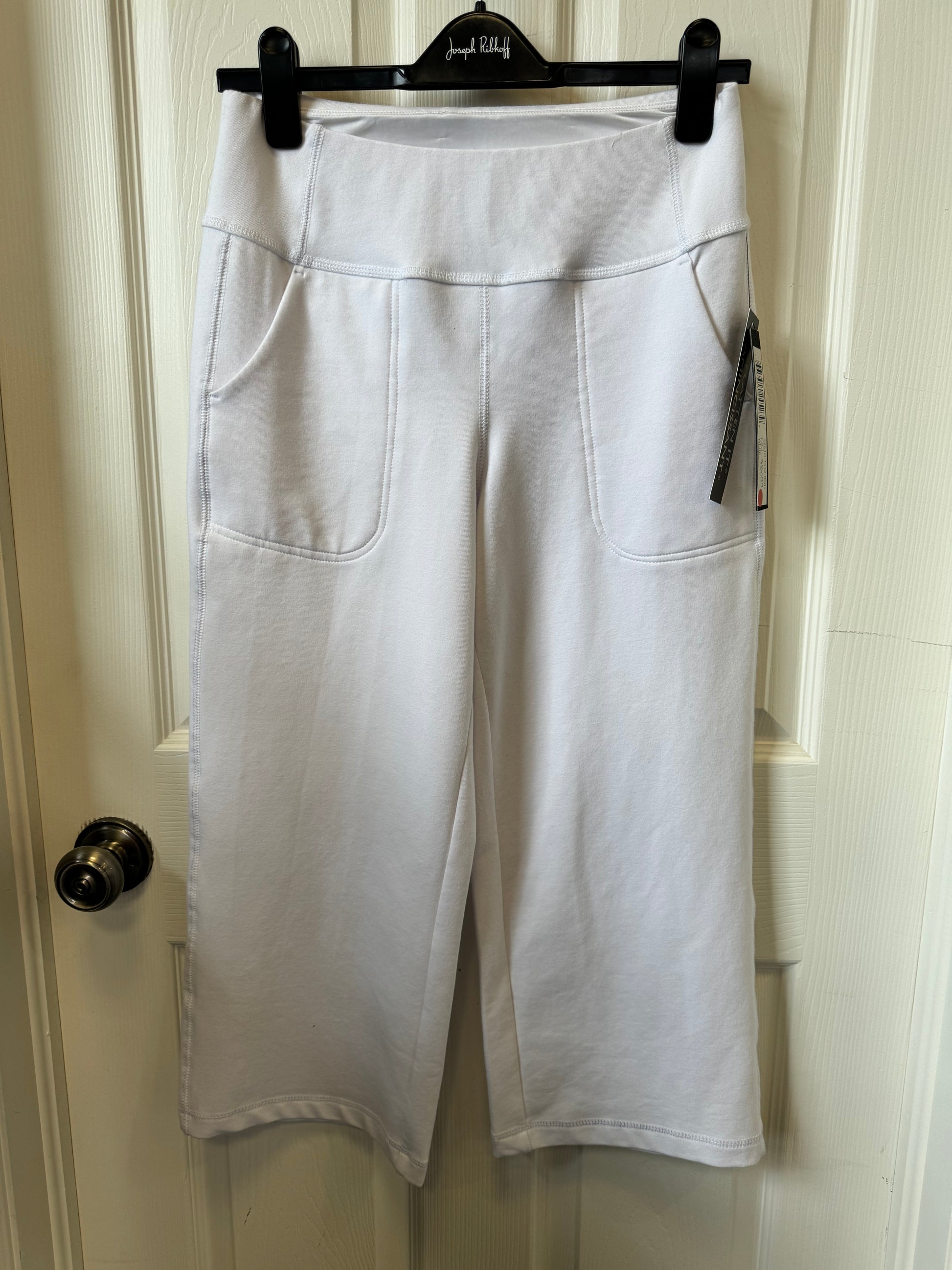 A pair of Tribal White Jogger Pants - 7264O-4532-0001 hangs on a black hanger against a white door. These stylish and functional pants from the Tribal brand feature a high waistband and two large front pockets with a tag attached to one of them. The door has a metallic doorknob on the left side.