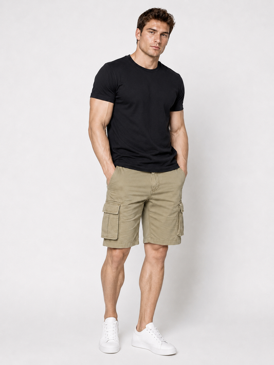 A man stands against a plain background, wearing a black t-shirt, white sneakers, and Silver Jeans Co. Collin Classic Fit Cargo Bermudas in Light Olive, with his hands in his pockets.