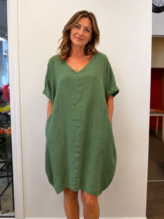 A woman with wavy brown hair smiles indoors wearing the ETERNELLE Linen Dress Green 6-SS26219-9, a loose, short-sleeved dress with pockets. She stands against a white wall with flowers and furniture in the background.