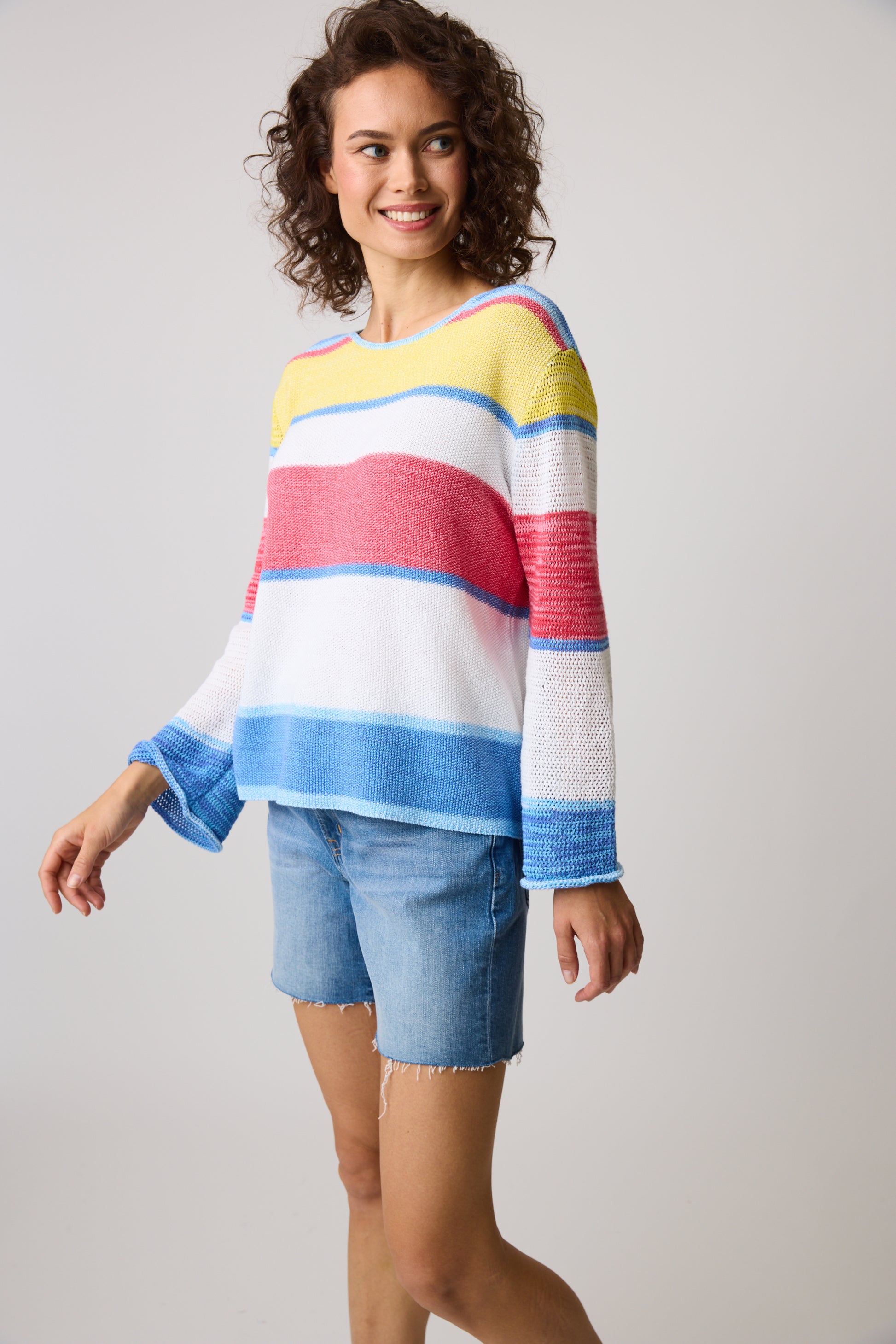 A person with curly hair smiles in a Parkhurst Cotton Country Naples Stripes Denim Combo 80117 sweater featuring yellow, blue, red, and white colors paired with denim shorts against a plain gray background.