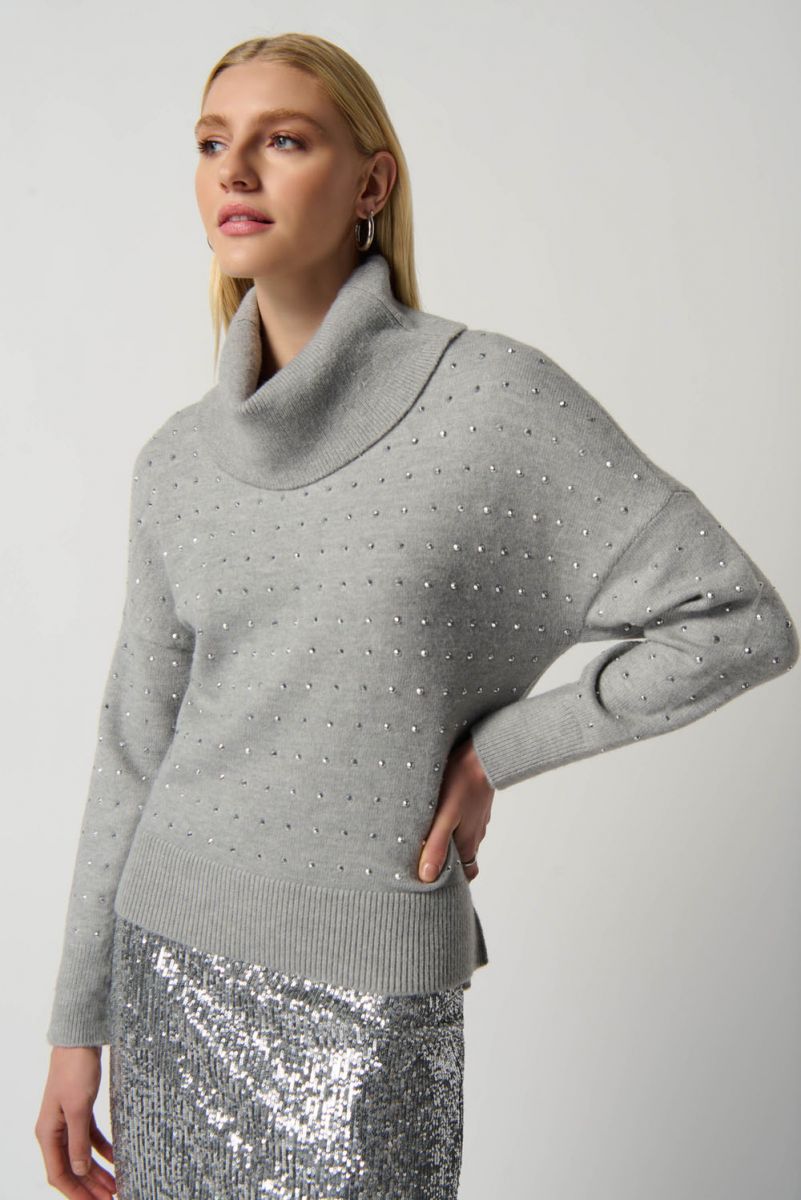 A person with long blonde hair is posing against a plain backdrop, wearing a cozy Light Grey Mélange Cowl Neck Sweater Style 234909 by Joseph Ribkoff adorned with small beads and a sparkling silver sequined skirt. They have their arm bent, with a hand resting on their hip, and are looking slightly to the side.