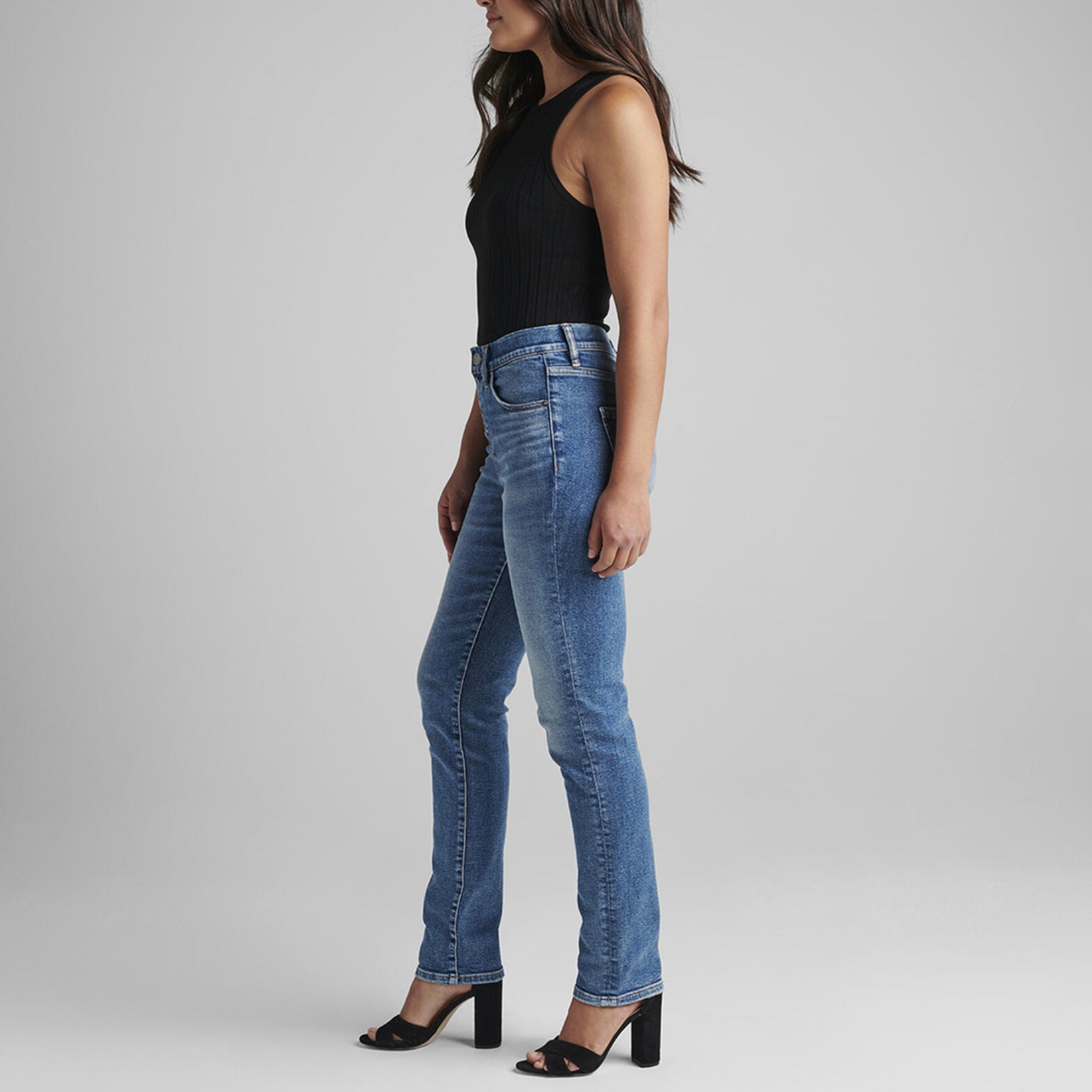 A person stands against a plain gray background, wearing a black sleeveless top and the Valentina High Rise Straight Leg Pull-On Jeans from JAG. They have long, wavy dark brown hair, and are wearing black open-toe heels with their arms relaxed by their sides.