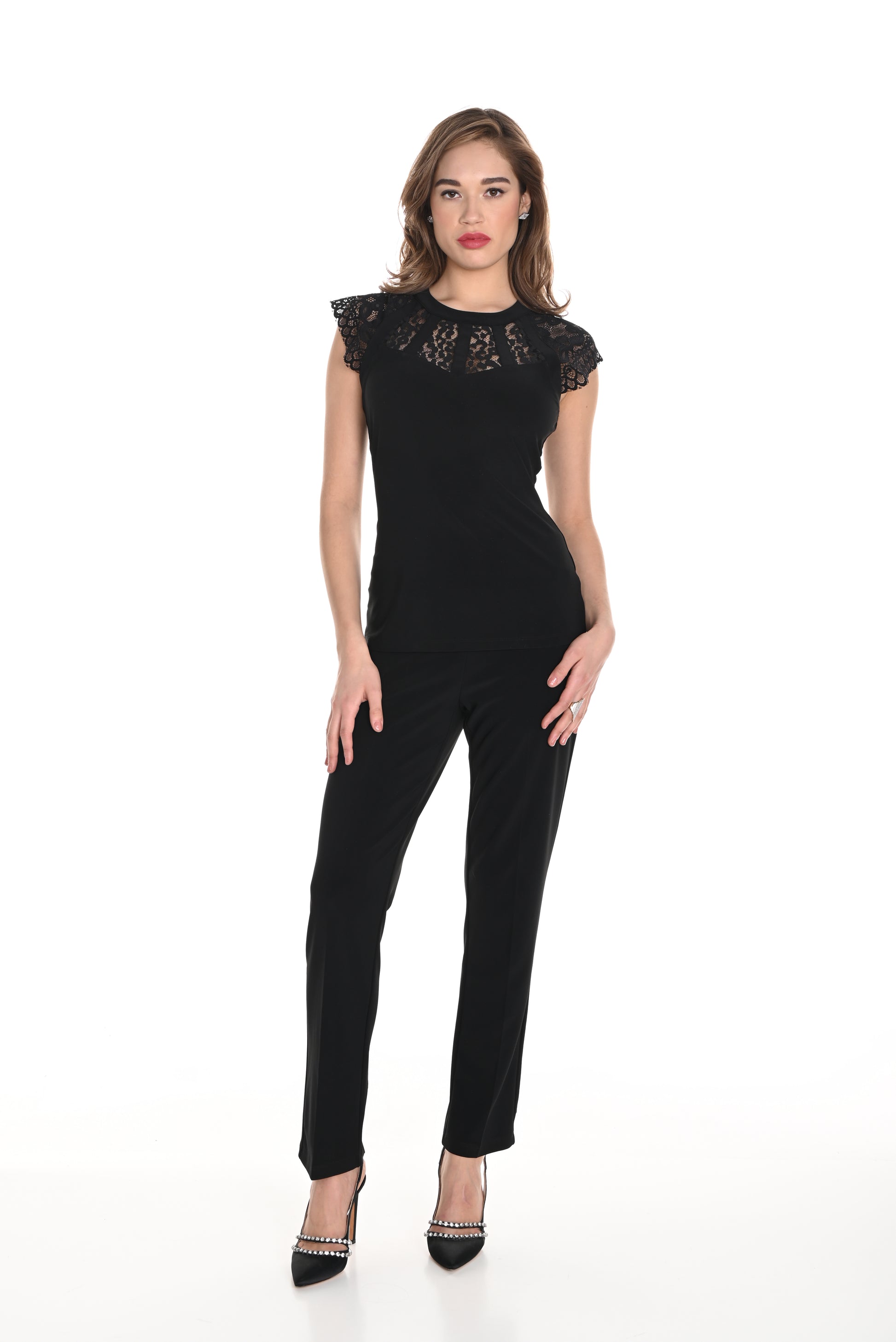 A woman with light brown, loosely styled hair stands against a white background, wearing a black lace top and Frank Lyman’s Bow Detail Knit Pant in black (Style 244019), paired with black heels featuring silver studs. She has a neutral expression on her face.