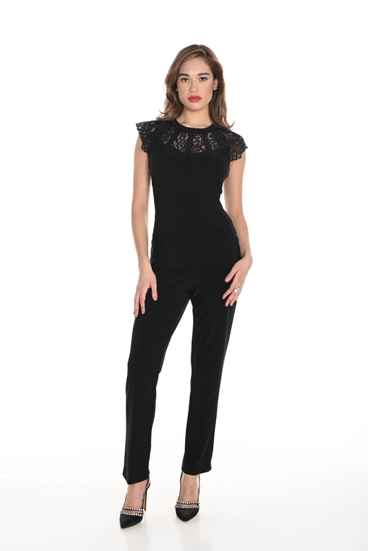 A woman with light brown, loosely styled hair stands against a white background, wearing a black lace top and Frank Lyman’s Bow Detail Knit Pant in black (Style 244019), paired with black heels featuring silver studs. She has a neutral expression on her face.