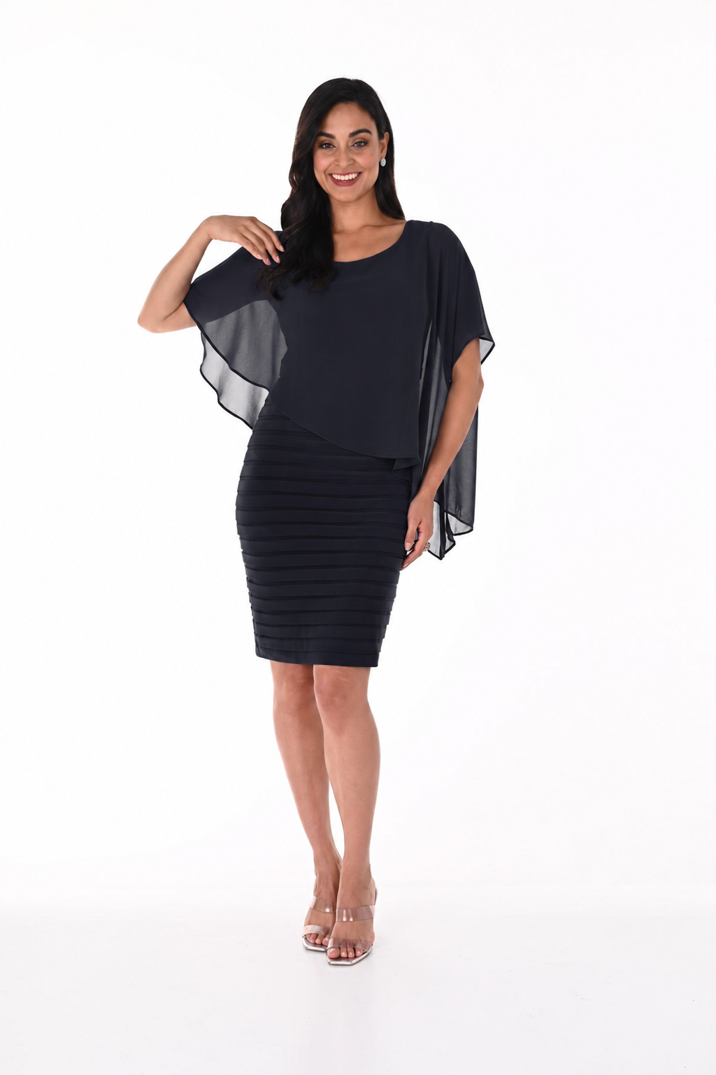 A woman stands and smiles, wearing a short, fitted navy Frank Lyman Chiffon Cape Dress (Style 51027) featuring a sheer chiffon overlay cape, paired with clear heeled sandals, set against a plain white background.