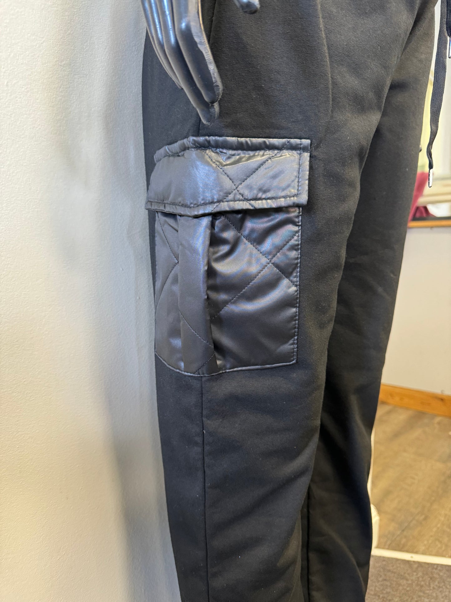 A mannequin dressed in ETERNELLE's Black Joggers with patch pockets, featuring a drawstring waist and elastic cuffs. The jogger pants, made in Italy, have stylish cargo pockets with a glossy zippered detail and are paired with a plain white top. The mannequin's hands are visible, resting by its sides.