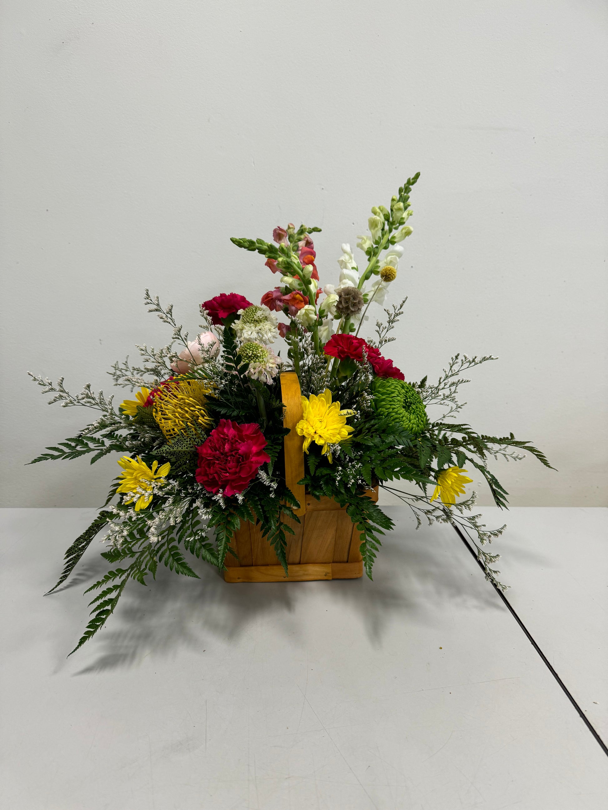 The "Pop of Colour - Basket" by Posies Flowers & Fashion features a wooden container brimming with a vibrant assortment of specific flowers, including red carnations, white snapdragons, yellow chrysanthemums, and assorted greenery, elegantly arranged on a white surface against a plain background.