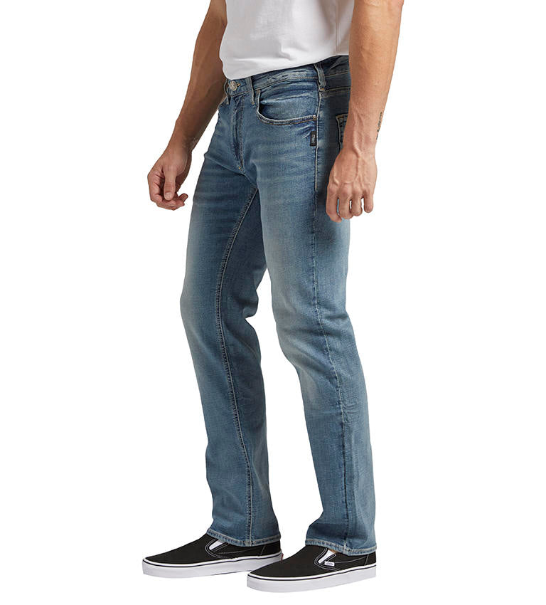 A person is standing sideways, dressed in Allan Slim Fit Straight Leg Jeans from Silver Jeans Co., a white T-shirt, and black slip-on shoes. The jeans have a fitted silhouette with slight fading on the thighs and knees. The person's arm is relaxed by their side.