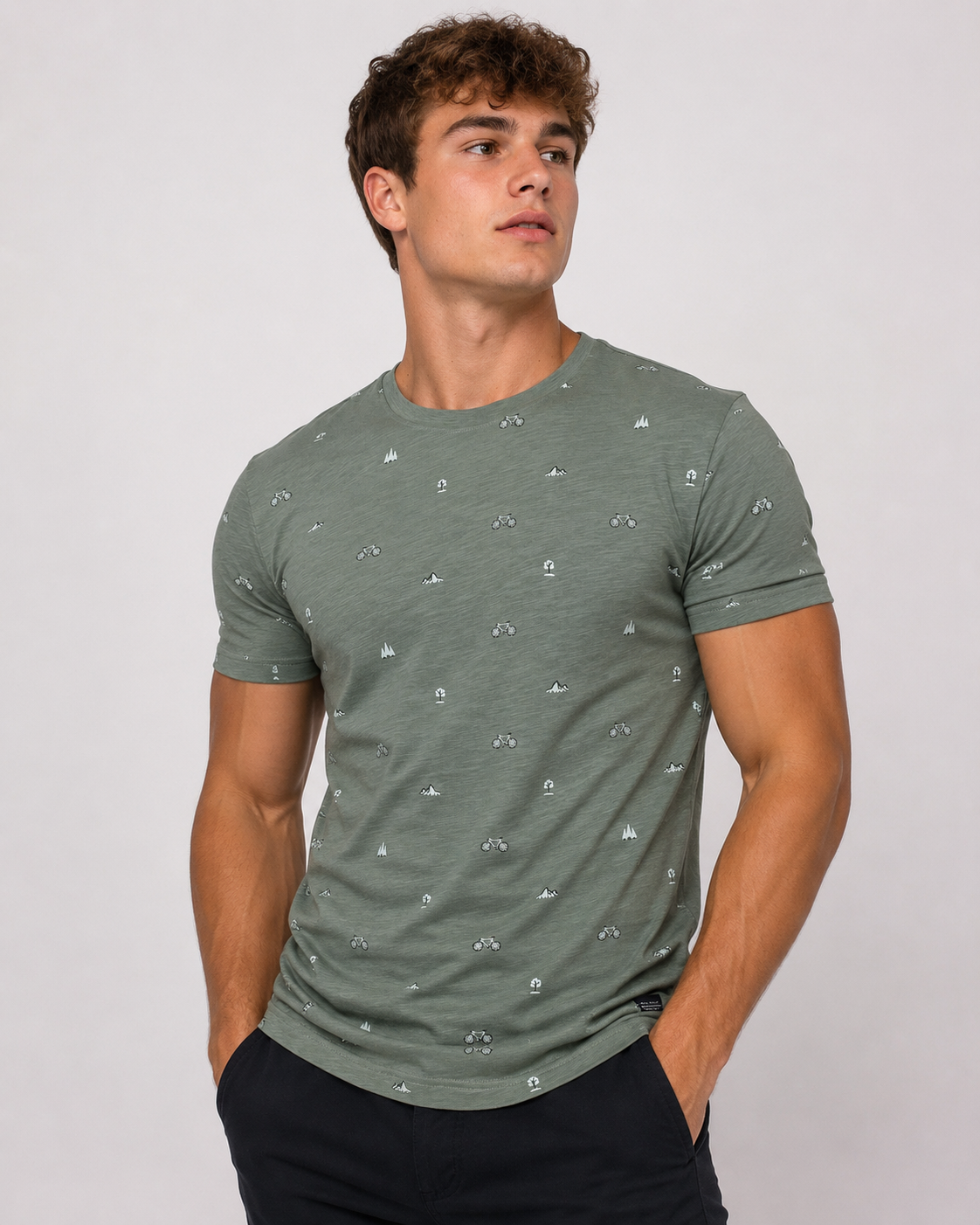 A young man with curly brown hair wears the Men’s Cotton T-Shirt - GREEN from Silver Jeans Co., featuring subtle white and black patterns, paired with dark pants. He stands with hands in pockets, looking to the side against a light background.