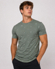 A young man with curly brown hair wears the Men’s Cotton T-Shirt - GREEN from Silver Jeans Co., featuring subtle white and black patterns, paired with dark pants. He stands with hands in pockets, looking to the side against a light background.