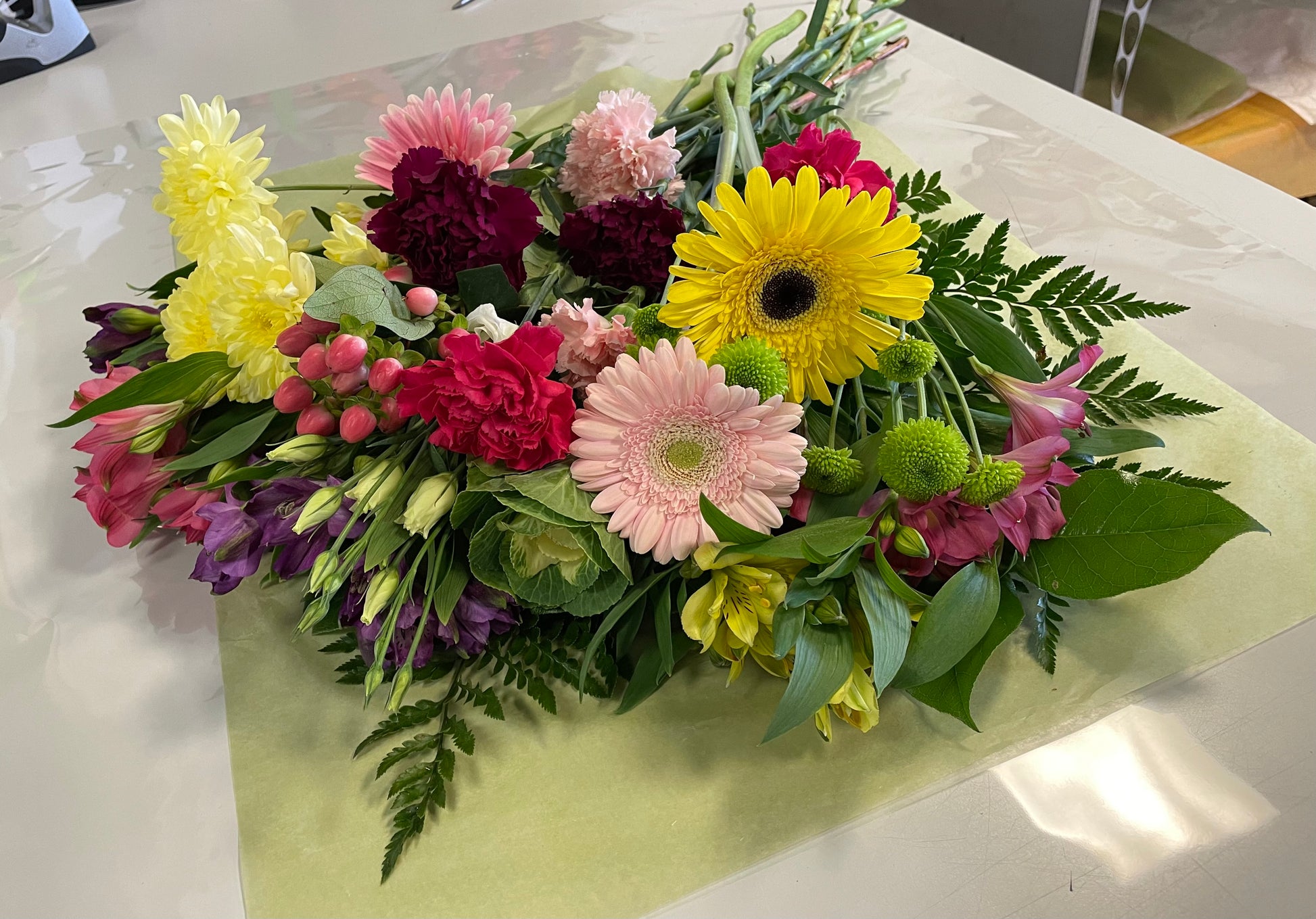 A stunning bouquet called Designer’s Choice Freshcuts from Posies Flowers & Fashion is resting on a white surface. The arrangement features vibrant orange and pink roses, cheerful yellow chrysanthemums, elegant white flowers, and assorted green foliage. It is partially wrapped in brown paper and elegantly tied with a burgundy ribbon.