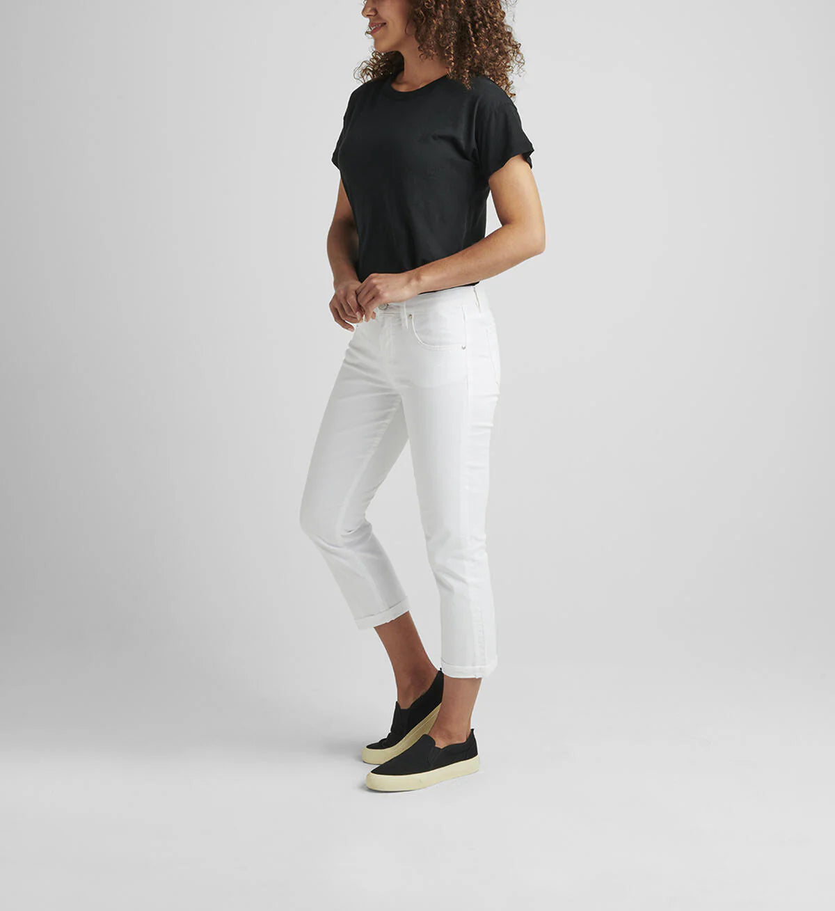 A person with curly hair, wearing a black t-shirt and the Cecilia Mid Rise Capri White from JAG, paired with black sneakers with white soles, is standing and facing away from the camera. The background is plain and gray.