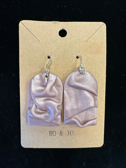 A pair of metallic pink, arched earrings with a draped fabric-like texture, crafted from polymer clay, is displayed on a beige card labeled "RO&JO" at the bottom and featuring a hole for hanging at the top. The product is named Ro&Jo Earrings 432. The background is black.