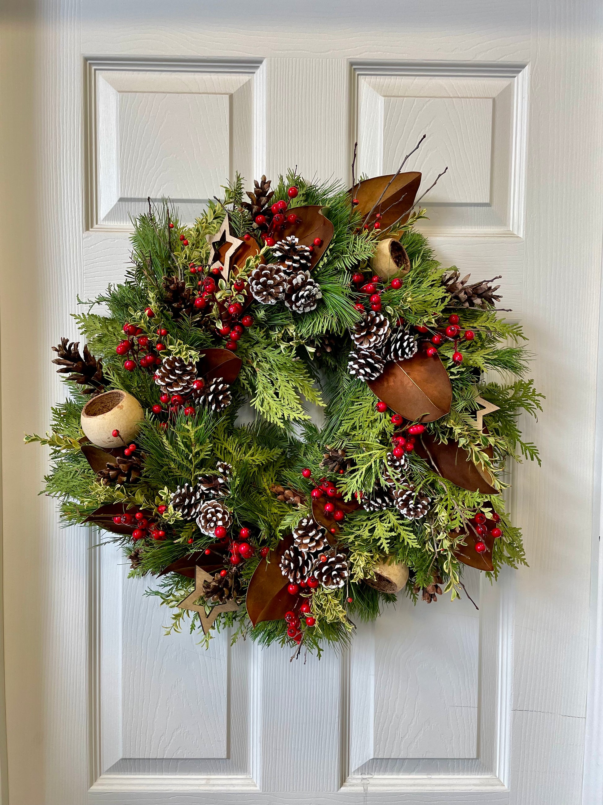 A Holiday Cheer Wreath from Posies Flowers & Fashion adorns the white door, featuring pinecones, red berries, magnolia leaves, and star-shaped ornaments. The fresh greens bring forth the holiday spirit, achieving a timeless festive appearance.