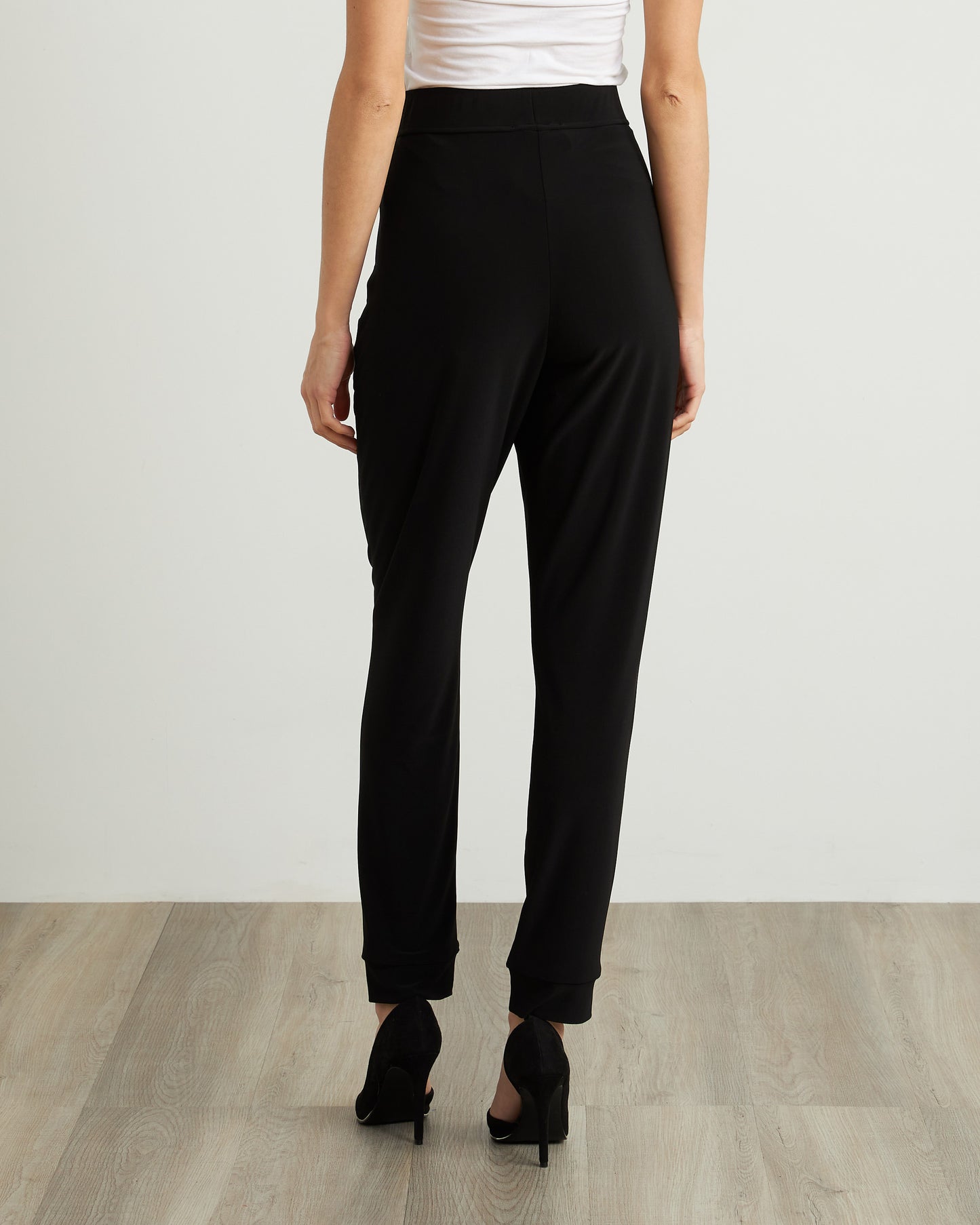 An individual wearing Joseph Ribkoff's Drawstring Waist Pants Style 211317, featuring zipper detailing in black, paired with a white top and black heels. The person stands on a wooden floor against a plain, light-colored background, seamlessly combining comfort and style.