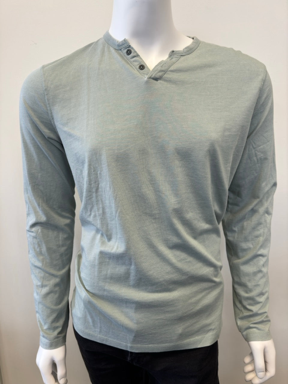 A mannequin dressed in the Silver Jeans Co. Shirt Long Sleeve - SAGE, featuring a classic 3-button design along the V-neckline. The sage green shirt, made from a comfortable cotton-polyester blend, is paired with black pants. The background is plain and white.