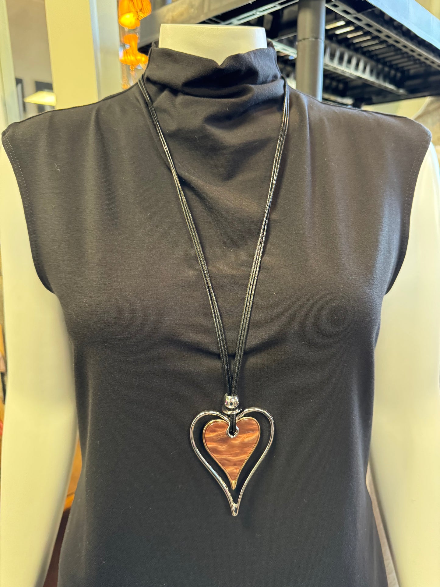 A mannequin highlights a sleeveless black top complemented by Necklace 11 from Elise Accessories. This elegant piece, with its metallic frame surrounding a reddish-brown heart-shaped center, takes center stage in this chic retail display.