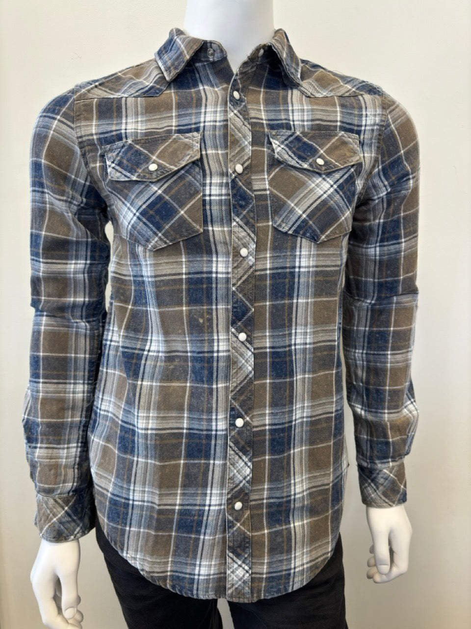 A mannequin wears the Shirt Long Sleeve - OLIVE by Silver Jeans Co., showcasing a plaid design in shades of brown, blue, and white. Crafted from 100% cotton, this button-up shirt features two front chest pockets with buttoned flaps. The mannequin's hand is partially visible on the right side as it stands against a plain, light-colored background.