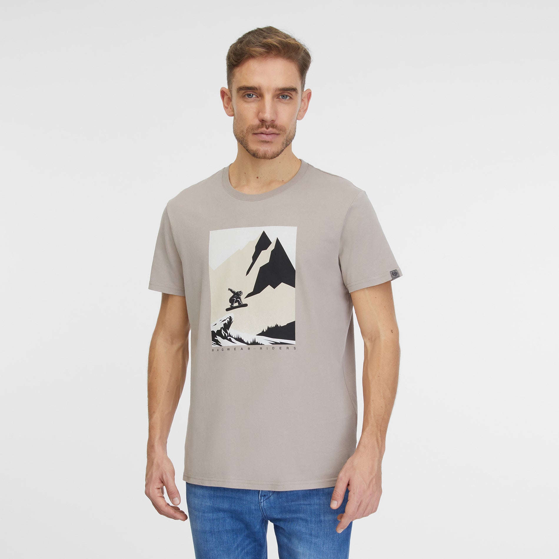 A man with short hair and stubble is wearing the Roggero T-Shirt 2422-15024-7001 from RAGWEAR, a beige, eco-friendly shirt featuring a mountain and bear graphic on the front. He pairs it with blue jeans and stands against a plain white background, looking directly at the camera with a neutral expression.