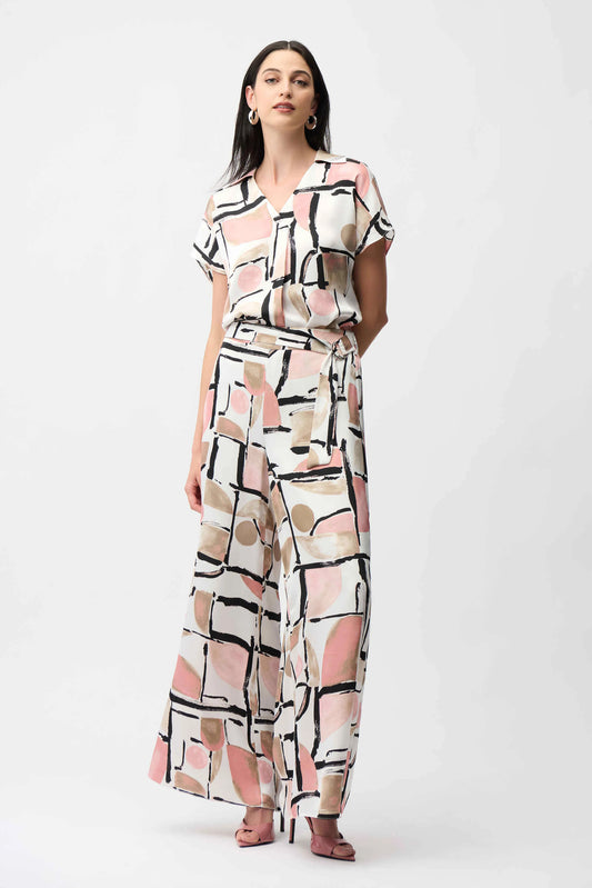 A woman wears the Joseph Ribkoff Recycled Satin Abstract Print Boxy Top with a V-neckline, styled as a matching set with wide-leg pants, pink heeled sandals, and hoop earrings.