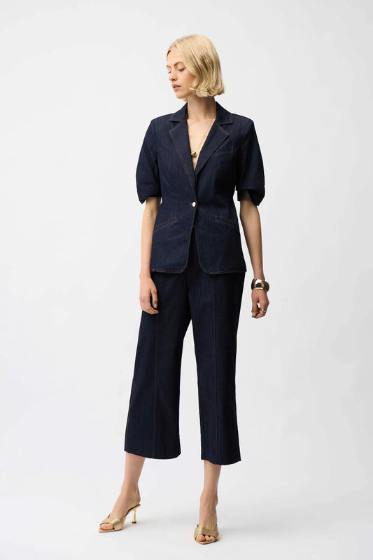 A woman with blonde hair stands against a plain background wearing the Joseph Ribkoff Denim Fitted Blazer 261968, featuring short puff sleeves and shank buttons, paired with cropped wide-leg pants, heeled sandals, and a bracelet.
