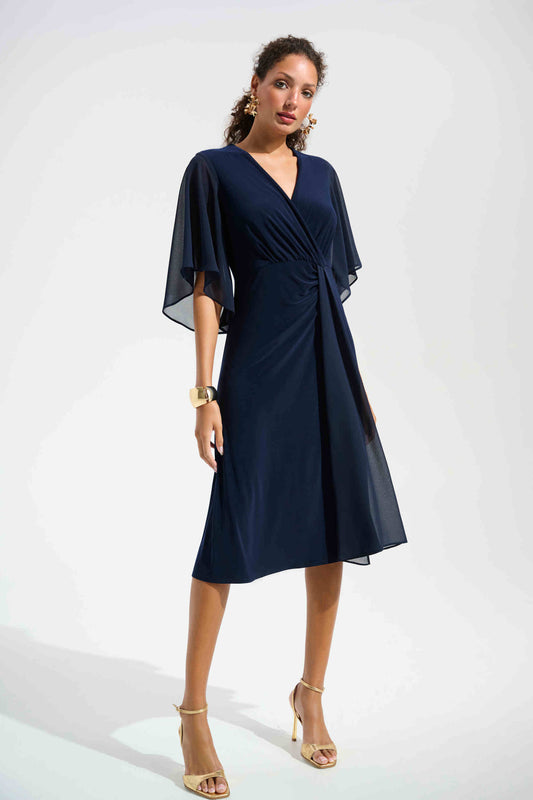 A woman wears the Joseph Ribkoff Signature Silky Knit Fit and Flare Dress With Chiffon Draped Sash in navy, featuring a V-neck and bell sleeves, paired with gold sandals, hoop earrings, and a chunky bracelet against a plain light background.