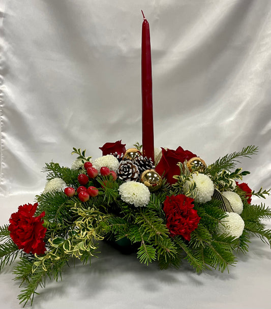 Join the Dec 18 Centrepiece Workshop at 6 PM by Posies Flowers & Fashion Inc. and craft a striking festive centerpiece. This arrangement includes a tall red candle surrounded by red and white flowers, pine cones, gold ornaments, and lush green foliage, all beautifully set against a crisp white background to enhance any holiday ambiance.