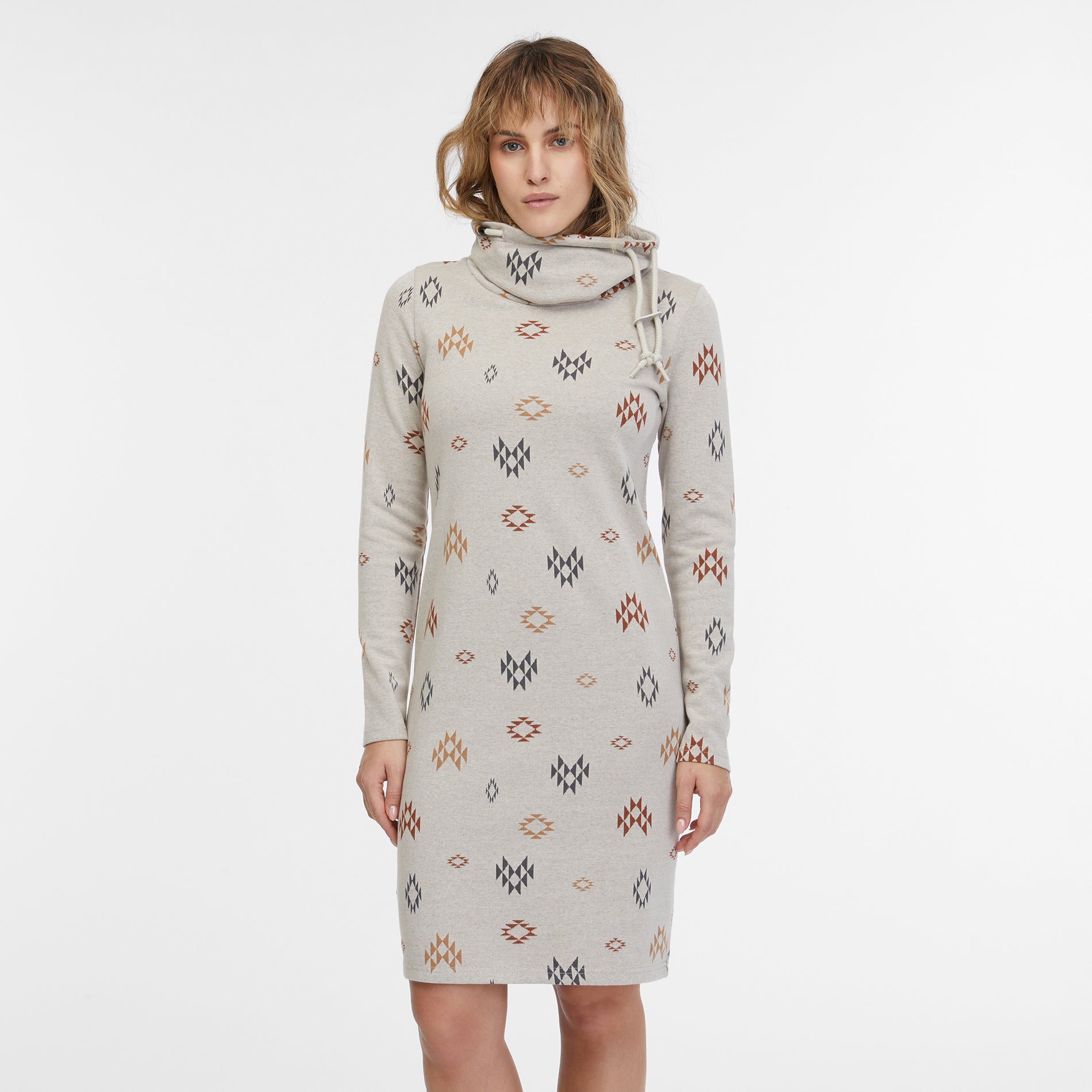 A person with shoulder-length hair is wearing the Sweater Dress Chloenette Ecru by RAGWEAR, a light-gray long-sleeve dress adorned with a geometric diamond pattern in various colors. The dress features a high, loose neck with a drawstring. The background is plain white.