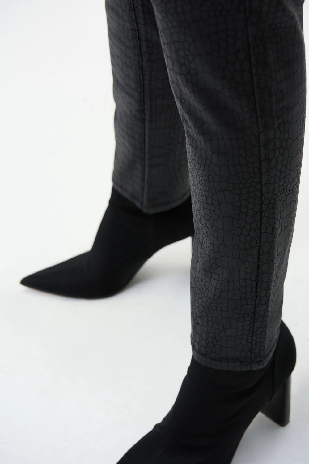 A person is standing against a plain white background, wearing Joseph Ribkoff's Black Croc Print Pants Style 224943 and sleek black ankle boots. Only the midsection and legs are visible, with hands resting at the sides.