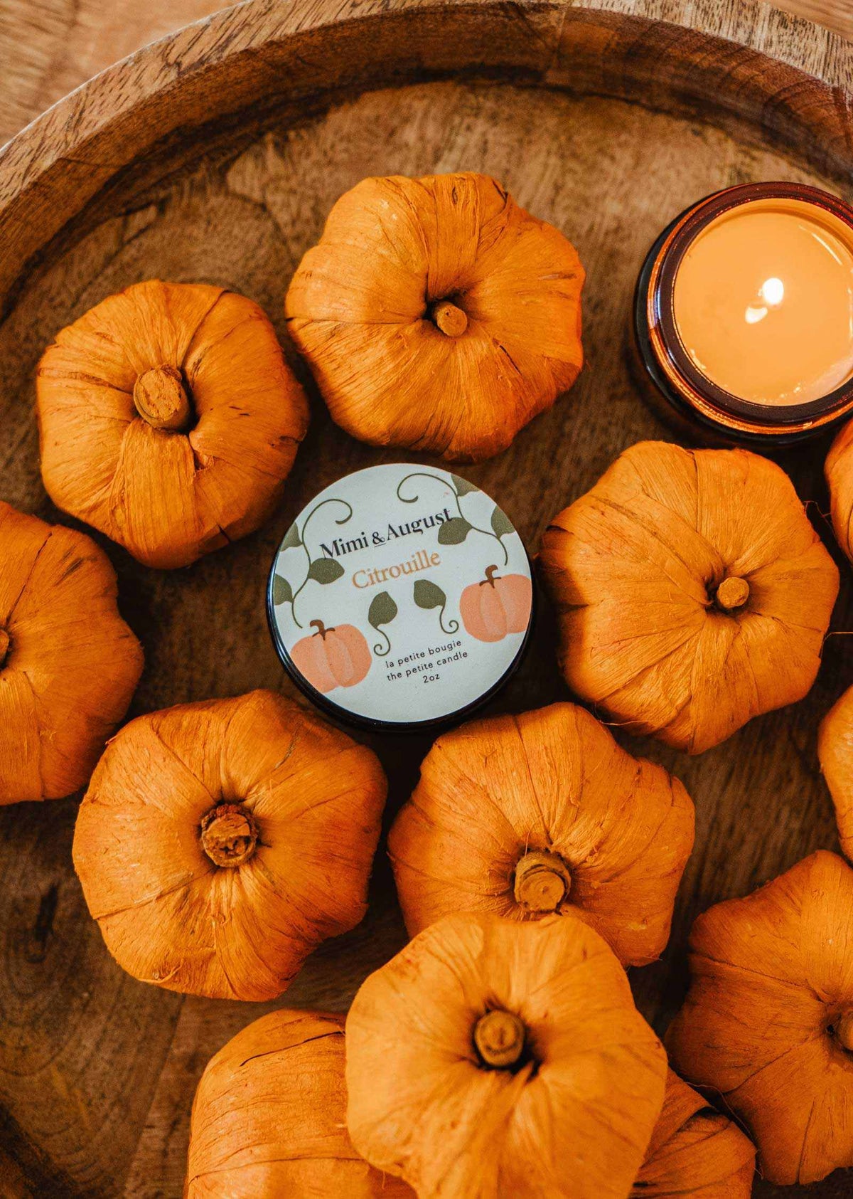 A round wooden tray with small orange pumpkins surrounds a Mimi & August Citrouille – 2 oz Soy Candle tin and a lit orange soy wax candle in a glass holder, creating the perfect fall centerpiece.