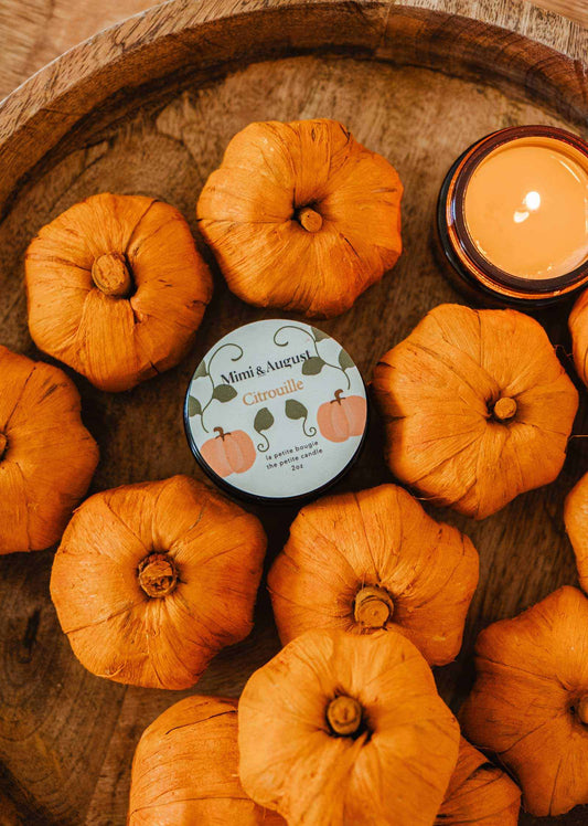 A round wooden tray with small orange pumpkins surrounds a Mimi & August Citrouille – 2 oz Soy Candle tin and a lit orange soy wax candle in a glass holder, creating the perfect fall centerpiece.