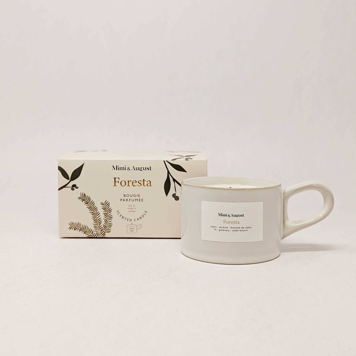 A white candle from Mimi & August, nestled in a reusable 5.5 oz mug-shaped holder, sits beside its cream-colored Foresta-scented box adorned with botanical illustrations and minimalist design, evoking the fresh aroma of a true Christmas tree forest experience.