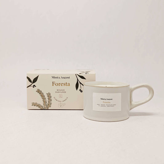 A white candle from Mimi & August, nestled in a reusable 5.5 oz mug-shaped holder, sits beside its cream-colored Foresta-scented box adorned with botanical illustrations and minimalist design, evoking the fresh aroma of a true Christmas tree forest experience.