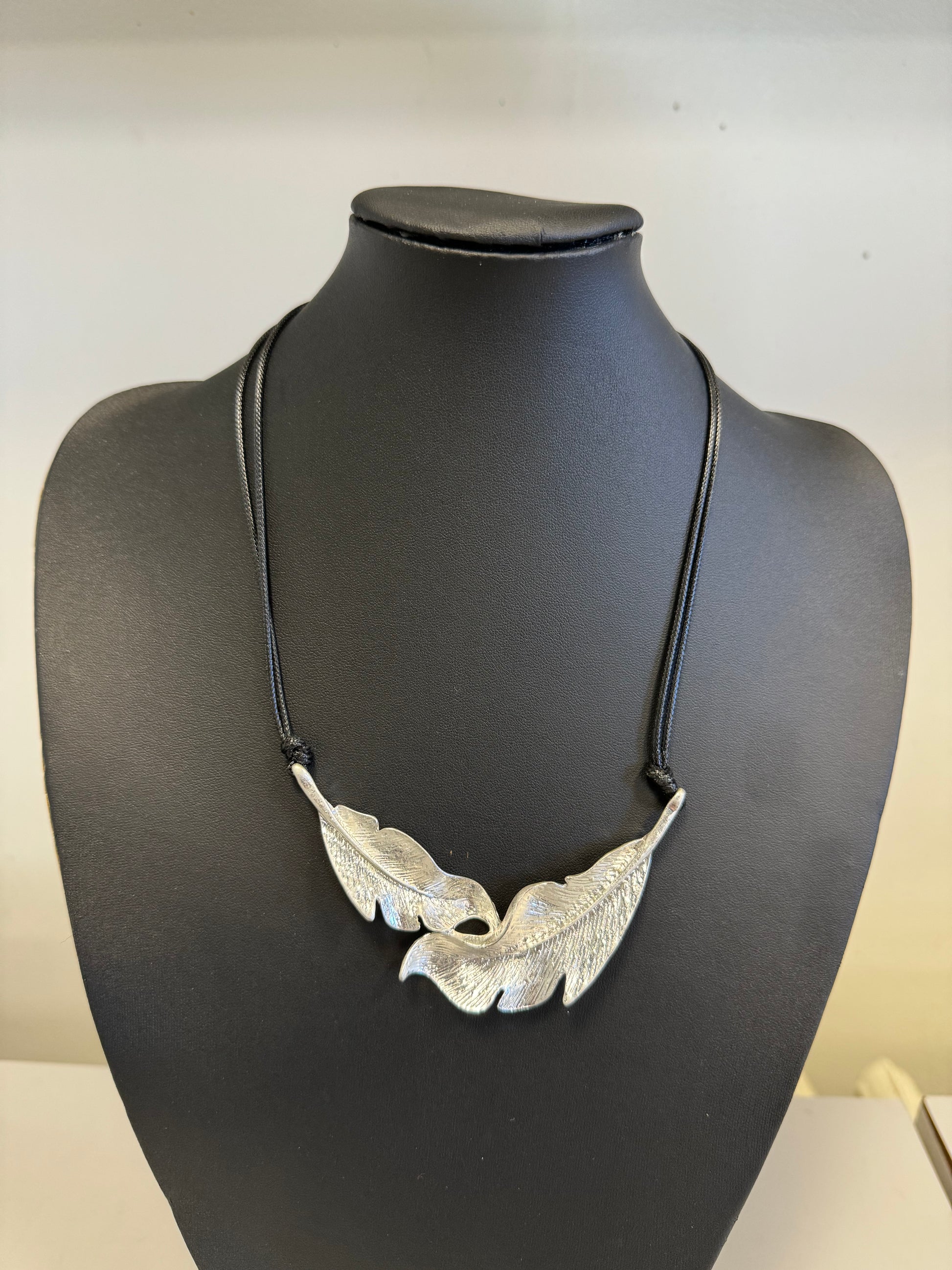 The Roxanne 32 Necklace from Elise Accessories, an exquisite piece from their collection, showcases two intricately designed feather pendants hanging on a black cord. It is beautifully presented on a black bust, with a plain light-colored background that enhances its elegant fashion appeal.