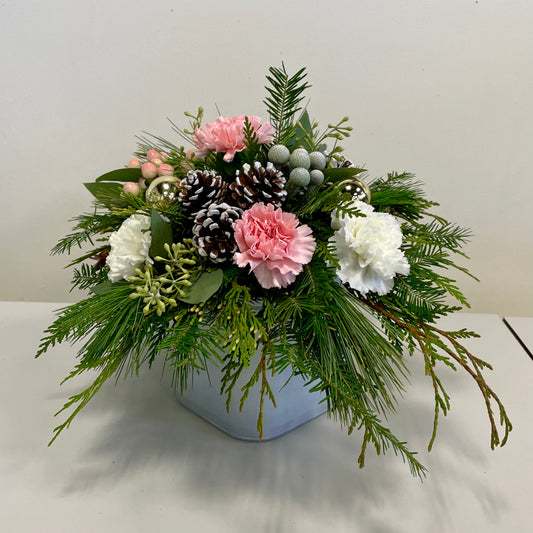 The Holiday Pink Centrepiece by Posies Flowers & Fashion is a festive table arrangement in a white vase, showcasing pink and white carnations, pinecones, and lush green foliage with pine branches and leaves. Small berry accents provide an additional touch of color and texture.