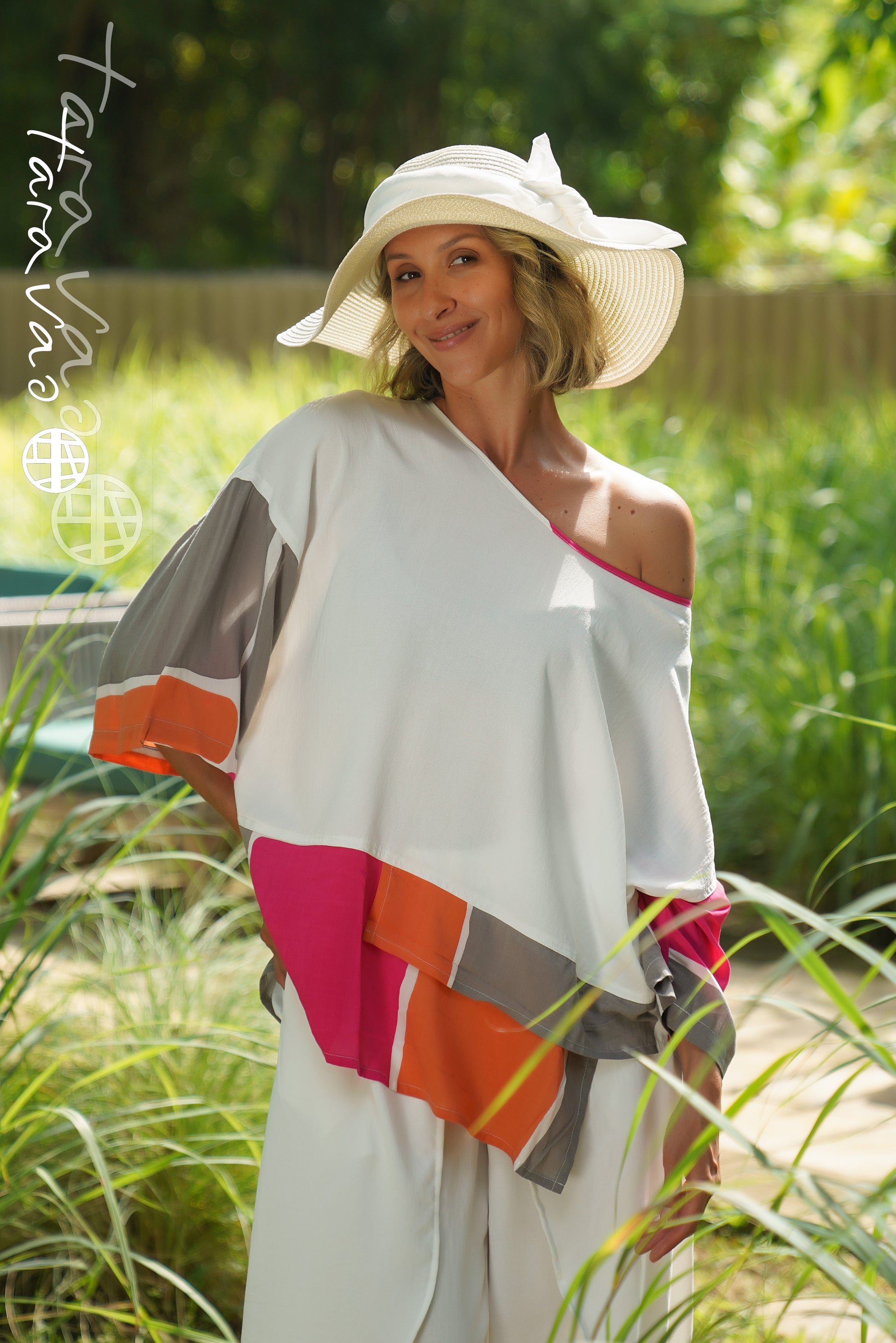 A woman wearing the TaraVao Top Maora 2137, a vibrant layered color-block top with an asymmetrical hem, smiles at the camera while standing outdoors in tall grass, sunlight highlighting her look. She pairs it with white pants and a wide-brimmed white hat.