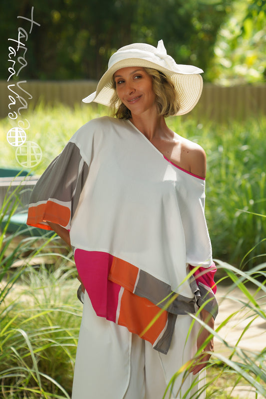 A woman wearing the TaraVao Top Maora 2137, a vibrant layered color-block top with an asymmetrical hem, smiles at the camera while standing outdoors in tall grass, sunlight highlighting her look. She pairs it with white pants and a wide-brimmed white hat.