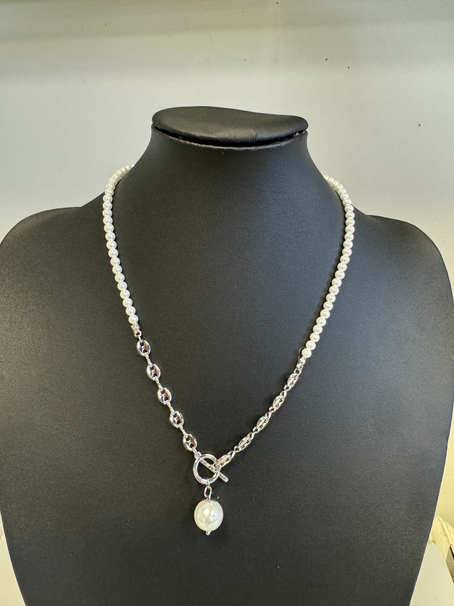 The Fiona 21 Necklace from Elise Accessories is showcased on a black stand, featuring a delicate strand of small white pearls intertwined with silver chain links, and accented by a large pearl pendant. The front is elegantly adorned with the clasp.