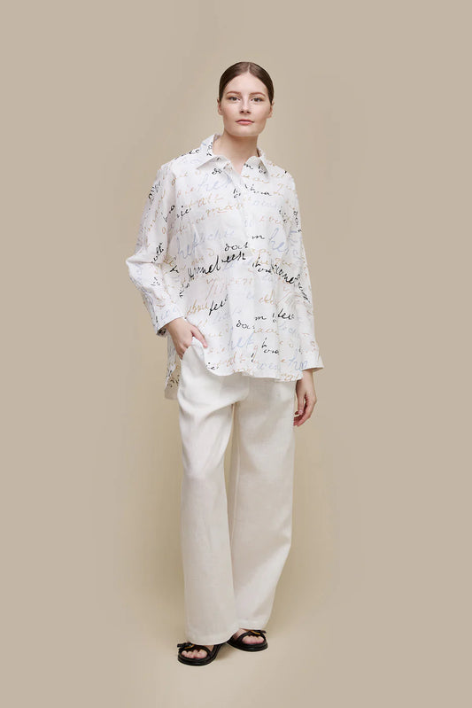 A person stands against a beige background, wearing the UCHUU Tunic Style Blouse (CS24-632) with handwritten-style text patterns. They pair it with loose white pants and black sandals. Their right hand rests in the pant pocket, and they have a neutral expression.