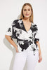 A woman with straight blonde hair exudes casual elegance, wearing a Vanilla/Black Wrap Front Top Style 232058 by Joseph Ribkoff, paired with white pants. She stands against a plain white background, one hand casually resting on her hip, looking confidently at the camera.