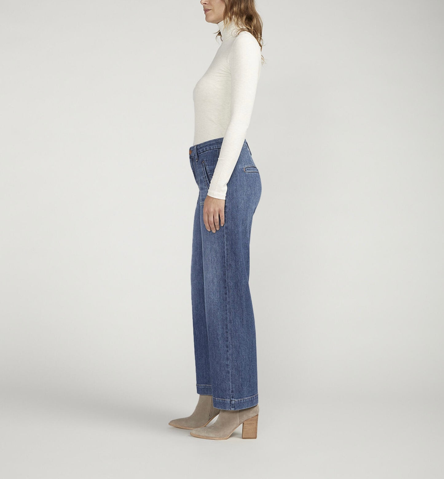 A person standing against a plain background is wearing a white turtleneck long-sleeve shirt, **JAG's Sophia High Rise Wide Leg Jeans in medium indigo wash with front pockets**, and light-colored ankle boots. Their hands are in their pockets and they are facing forward.